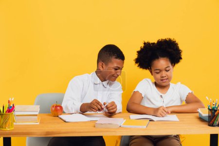 Foto de Feliz colegiala afroamericana y colegial de raza mixta sentados juntos en el escritorio y estudiando sobre fondo amarillo. Regreso al concepto escolar. - Imagen libre de derechos