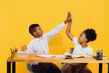 Photo for Happy african american schoolgirl and mixed race schoolboy sitting together at desk and giving high five to each other on yellow background. Back to school concept. - Royalty Free Image