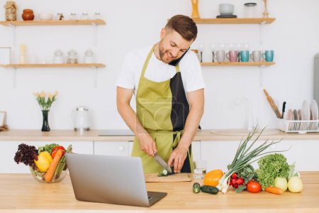 Téléchargez les photos : Jeune homme attrayant cuisine dans la cuisine avec un ordinateur portable sur la table, parler sur un smartphone. - en image libre de droit