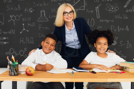 Photo for A female teacher poses with school children on the background of blackboard. International team. Back to school. - Royalty Free Image