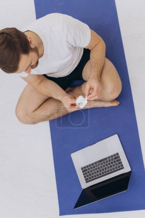 Photo for Attractive bearded man holding wireless headphones near laptop and starting morning exercise or yoga. - Royalty Free Image