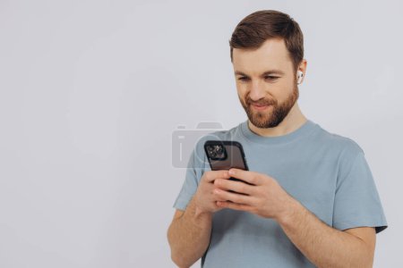 Téléchargez les photos : Portrait d'un homme moderne millénaire avec une barbe tenant un smartphone et écoutant quelque chose dans un casque sans fil sur un fond blanc avec espace de copie - en image libre de droit