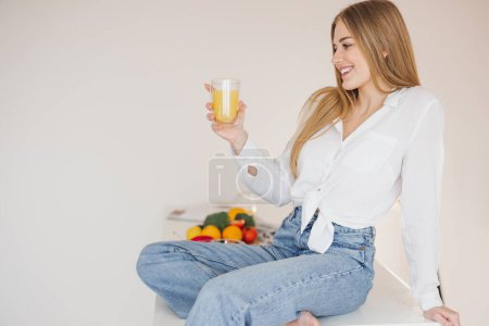 Téléchargez les photos : Femme blonde heureuse et souriante assise sur la table de cuisine et buvant du jus d'orange parmi les ingrédients de cuisine. - en image libre de droit
