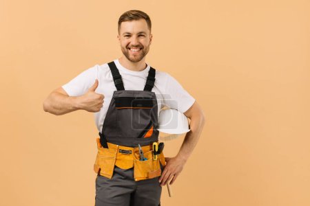 Téléchargez les photos : Réparateur masculin positif à la maison tenant un casque et montrant parfaitement sur un fond beige - en image libre de droit