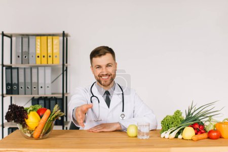 Téléchargez les photos : Heureux médecin nutritionniste assis sur le lieu de travail au bureau parmi les légumes frais et serrant la main du patient, concept de plan de régime - en image libre de droit