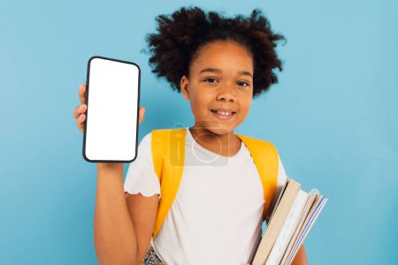 Foto de Emocionado Little African American School Girl sosteniendo una gran celda en la mano presentando la pantalla vacía del teléfono en el fondo del estudio azul. Smartphone Display Mockup - Imagen libre de derechos