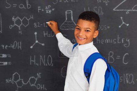 Photo for Happy mixed race schoolboy solving problems near the blackboard at school, back to school concept. - Royalty Free Image