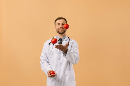 Photo for The male nutritionist doctor with stethoscope smiling and juggling tomatoes on beige background, diet plan concept - Royalty Free Image