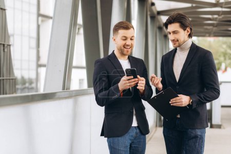 Téléchargez les photos : Deux hommes d'affaires modernes discutent de quelque chose et se montrent sur un smartphone dans le contexte des bureaux et des bâtiments urbains - en image libre de droit
