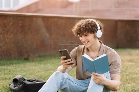 Photo for Attractive male student in headphones sitting on grass doing homework near university on sunny day - Royalty Free Image