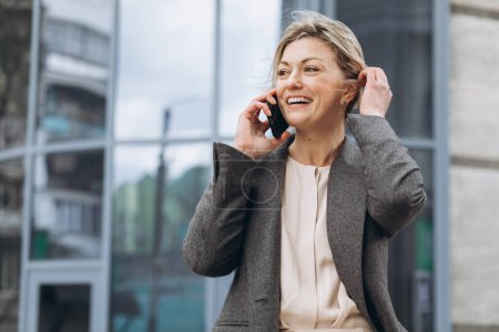 Foto de Retrato de una hermosa mujer de negocios madura en traje y chaqueta gris sonriendo y hablando por teléfono en los modernos edificios urbanos y de oficinas de fondo - Imagen libre de derechos