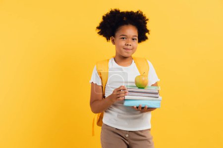 Photo for African american schoolgirl holding notebooks and books and showing something on yellow background, copy space. - Royalty Free Image