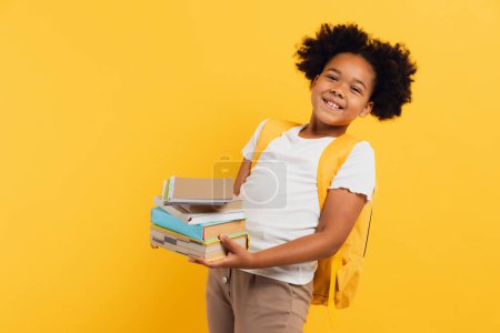 Téléchargez les photos : Heureuse écolière afro-américaine tenant des cahiers et des livres sur fond jaune, espace de copie. - en image libre de droit