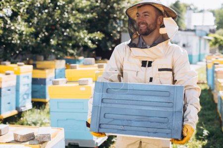 Foto de Feliz apicultor masculino en un traje protector mantiene una colmena en el colmenar - Imagen libre de derechos