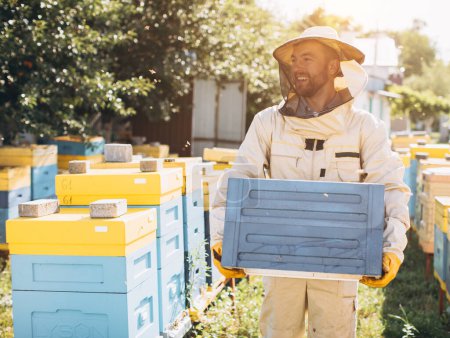 Foto de Feliz apicultor masculino en un traje protector mantiene una colmena en el colmenar - Imagen libre de derechos