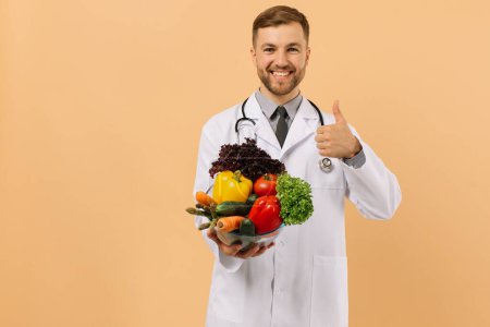 Foto de El nutricionista médico varón feliz con estetoscopio muestra verduras frescas en fondo beige, concepto de plan de dieta - Imagen libre de derechos