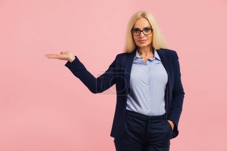Téléchargez les photos : Portrait de femme d'affaires attrayante d'âge moyen montrant quelque chose sur l'espace de copie sur fond rose - en image libre de droit