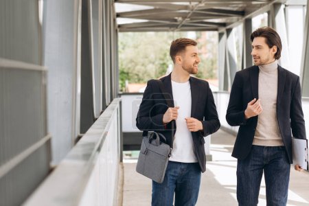 Téléchargez les photos : Deux hommes d'affaires heureux modernes marchant à travers un pont de la ville discutant de quelque chose dans le contexte des bureaux urbains et des bâtiments - en image libre de droit