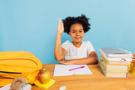 Foto de Feliz colegiala afroamericana sentada en el escritorio en clase y estirando la mano cuesta arriba sobre fondo azul. Regreso al concepto escolar. - Imagen libre de derechos