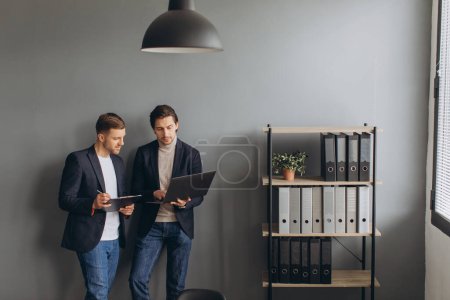 Téléchargez les photos : Jeunes hommes d'affaires debout ensemble tenant un ordinateur portable dans le bureau moderne, discuter - en image libre de droit