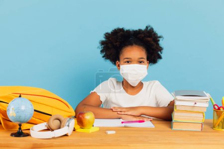 Photo for African American schoolgirl in a mask sits at a desk in class on a blue background. Back to school and covid-19 concept - Royalty Free Image