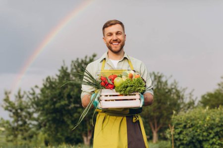 Photo for Happy gardener man holding basket with fresh vegetables on rainbow and garden background, gardening concept - Royalty Free Image