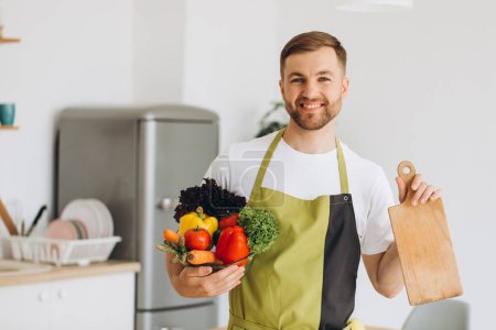 Photo for Portrait of happy man holding kitchen board and plate of fresh vegetables at home. - Royalty Free Image