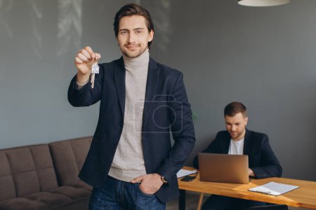 Photo for Smiling young modern man in a suit holds the keys to the bought apartment on the background of the realtor and office - Royalty Free Image