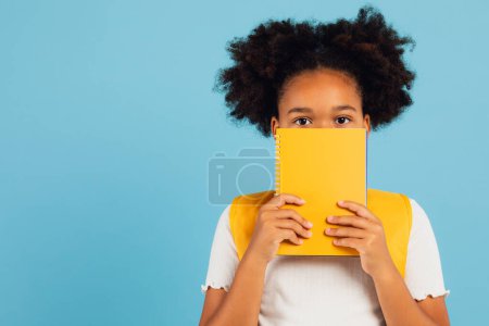 Téléchargez les photos : Drôle afro-américaine écolière couvrant son visage avec livre jaune sur fond bleu, concept de retour à l'école. - en image libre de droit