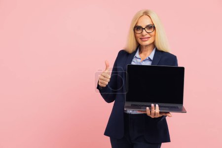 Téléchargez les photos : Belle femme d'affaires d'âge moyen dans des lunettes tenant un ordinateur portable et montrant la maquette de l'écran sur un fond rose - en image libre de droit