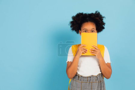 Foto de Divertida colegiala afroamericana cubriéndose la cara con libro amarillo sobre fondo azul, concepto de vuelta a la escuela. - Imagen libre de derechos