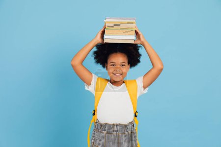 Téléchargez les photos : Happy African American écolière tenant des livres au-dessus de la tête sur fond bleu, concept de retour à l'école. - en image libre de droit