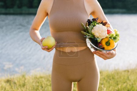 Téléchargez les photos : Femme végétalienne tenant pomme et assiette de légumes frais près du ventre en plein air - en image libre de droit