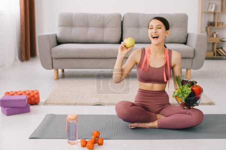 Photo for Young sporty woman sitting on yoga mat and holding variety of vegetables and apple, concept of weight loss and healthy eating. - Royalty Free Image