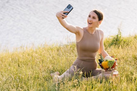 Foto de Mujer deportista feliz tomando selfie con plato de verduras frescas al aire libre - Imagen libre de derechos
