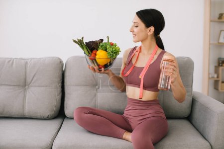 Photo for Young sporty woman sitting on sofa holding variety of vegetables, bottle of water and wrapped ruler, concept of weight loss and healthy eating. - Royalty Free Image