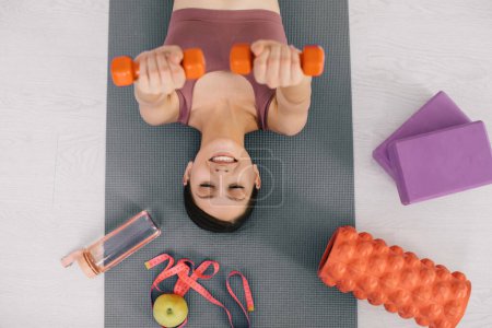 Photo for Attractive sporty young woman lying on yoga mat and around her sports equipment and apple, top view - Royalty Free Image