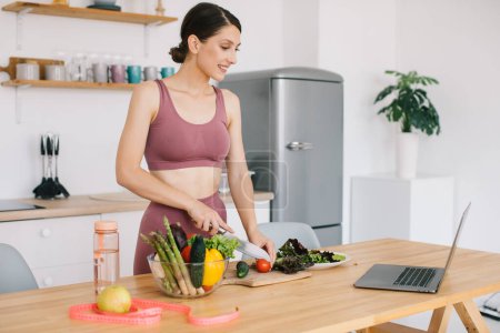 Téléchargez les photos : Heureuse femme sportive blogueuse nutritionniste préparer une salade avec des légumes frais et organise une vidéoconférence sur la saine alimentation sur ordinateur portable dans la cuisine - en image libre de droit