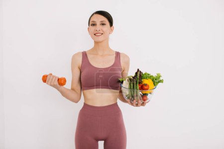 Téléchargez les photos : Belle jeune femme heureuse avec plateau de divers légumes sains et montrant ses muscles avec haltère isolé sur le fond blanc - en image libre de droit
