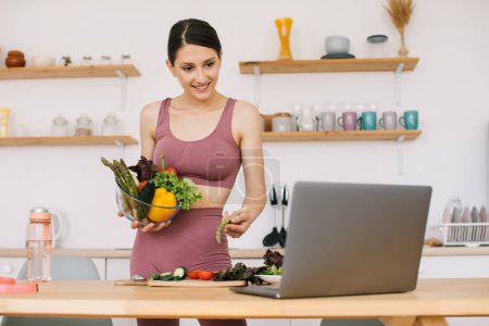 Téléchargez les photos : Heureuse femme sportive blogueuse nutritionniste préparer une salade avec des légumes frais et organise une vidéoconférence sur la saine alimentation sur ordinateur portable dans la cuisine - en image libre de droit