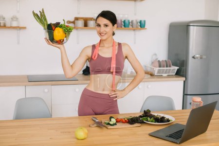 Foto de Retrato de una bloguera deportista feliz sosteniendo plato de verduras frescas y video conferencia líder sobre comer sano en la computadora portátil en la cocina - Imagen libre de derechos