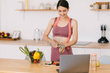 Téléchargez les photos : Portrait de blogueuse sportive heureuse faisant la cuisine aux asperges et vidéoconférence de premier plan sur le thème de la saine alimentation sur ordinateur portable dans la cuisine - en image libre de droit