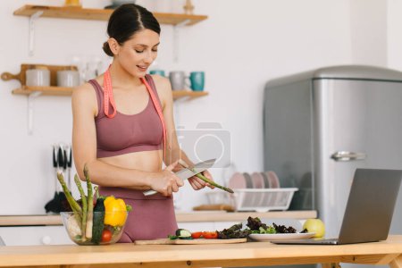 Téléchargez les photos : Portrait de blogueuse sportive heureuse faisant la cuisine aux asperges et vidéoconférence de premier plan sur le thème de la saine alimentation sur ordinateur portable dans la cuisine - en image libre de droit