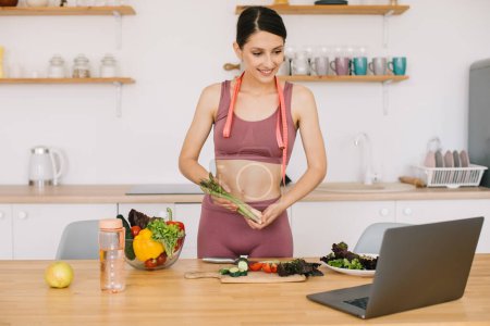 Photo for Happy sports woman blogger shows asparagus and leads a video conference on healthy eating on laptop in the kitchen - Royalty Free Image