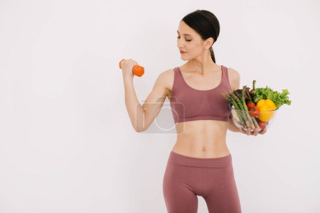 Photo for Beautiful happy young woman with tray of various healthy vegetables and showing her muscles with dumbbell isolated on the white background - Royalty Free Image