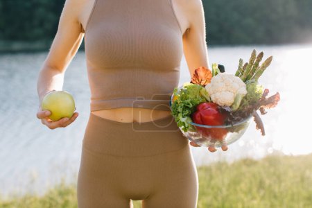 Foto de Mujer vegana sosteniendo manzana y plato de verduras frescas cerca del vientre al aire libre - Imagen libre de derechos