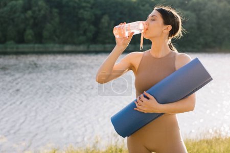 Foto de Mujer atlética bebe agua dulce y sostiene una esterilla de yoga al aire libre - Imagen libre de derechos
