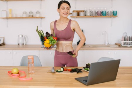 Photo for Athletic woman blogger nutritionist prepare a salad with fresh vegetables and conducts a video conference on healthy eating on laptop in the kitchen - Royalty Free Image