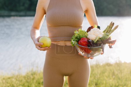 Téléchargez les photos : Femme végétalienne tenant pomme et assiette de légumes frais près du ventre en plein air - en image libre de droit