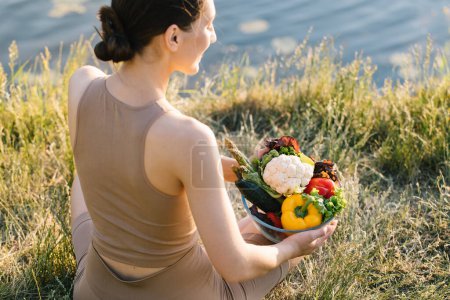 Foto de Deportiva mujer sosteniendo un plato de verduras frescas al aire libre junto al río, vista trasera - Imagen libre de derechos